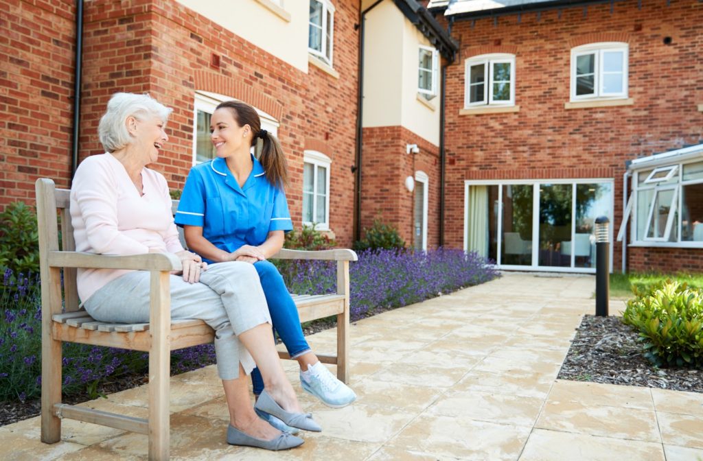 Senior woman accompanied by nurse as they sit on bench outside senior home community