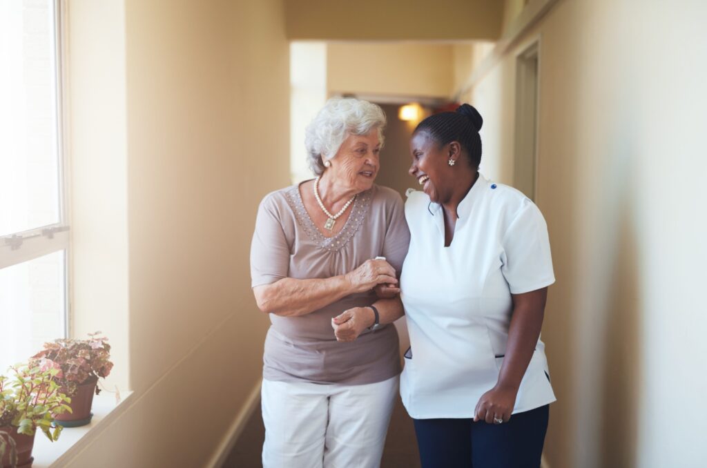 Senior walking arm-in-arm with a caregiver in a bright hallway, highlighting assisted living support and companionship.